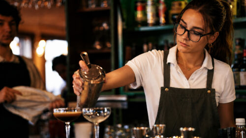 Bartender wearing glasses pours a cocktail from a shaker into a coupe glass at a bar A focused bartender in a white shirt and dark apron makes drinks behind a busy bar Photographer